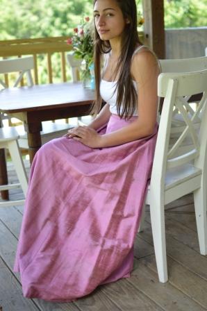 Young Brunette woman sitting at a dinning table outside with hands on her lap wearing a white spaghetti strapped shirt and 209 Walking skirt.