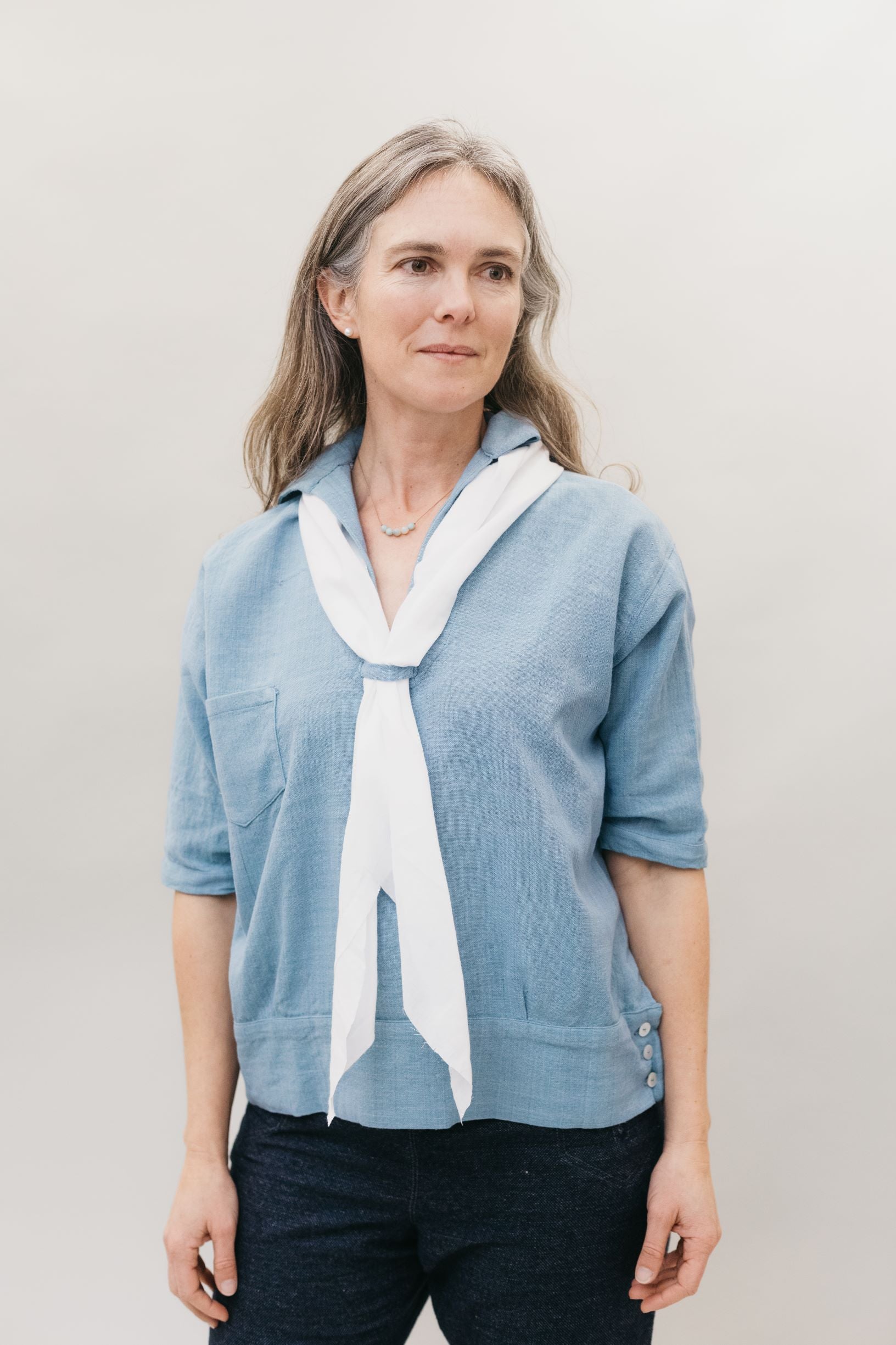A white woman standing in front of a studio white backdrop wearing the 211 Two middies v-neck with a contrasting scarf through the tab below the v-neck
