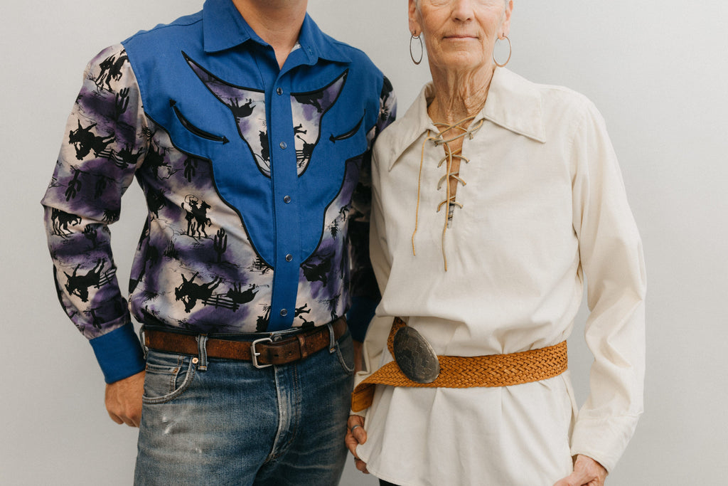 A man and a older woman standing in front of a white studio backdrop ,wearing 212 Five Frontier Shirts. Man wearing View D with decorative bull's head yoke and woman wearing View A with lace up front with belt around the waist.