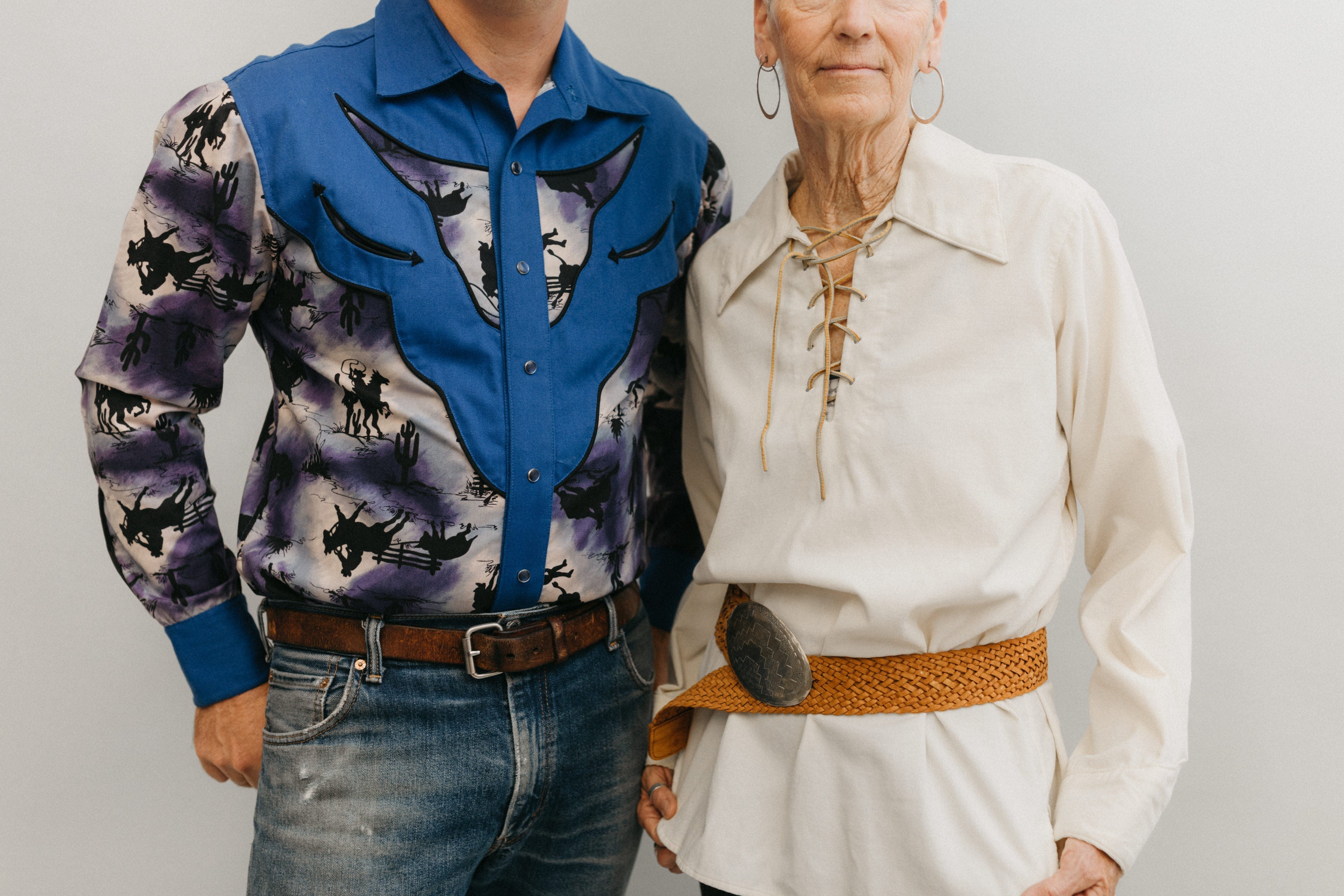 A man and a older woman standing in front of a white studio backdrop ,wearing 212 Five Frontier Shirts. Man wearing View D with decorative bull's head yoke and woman wearing View A with lace up front with belt around the waist.