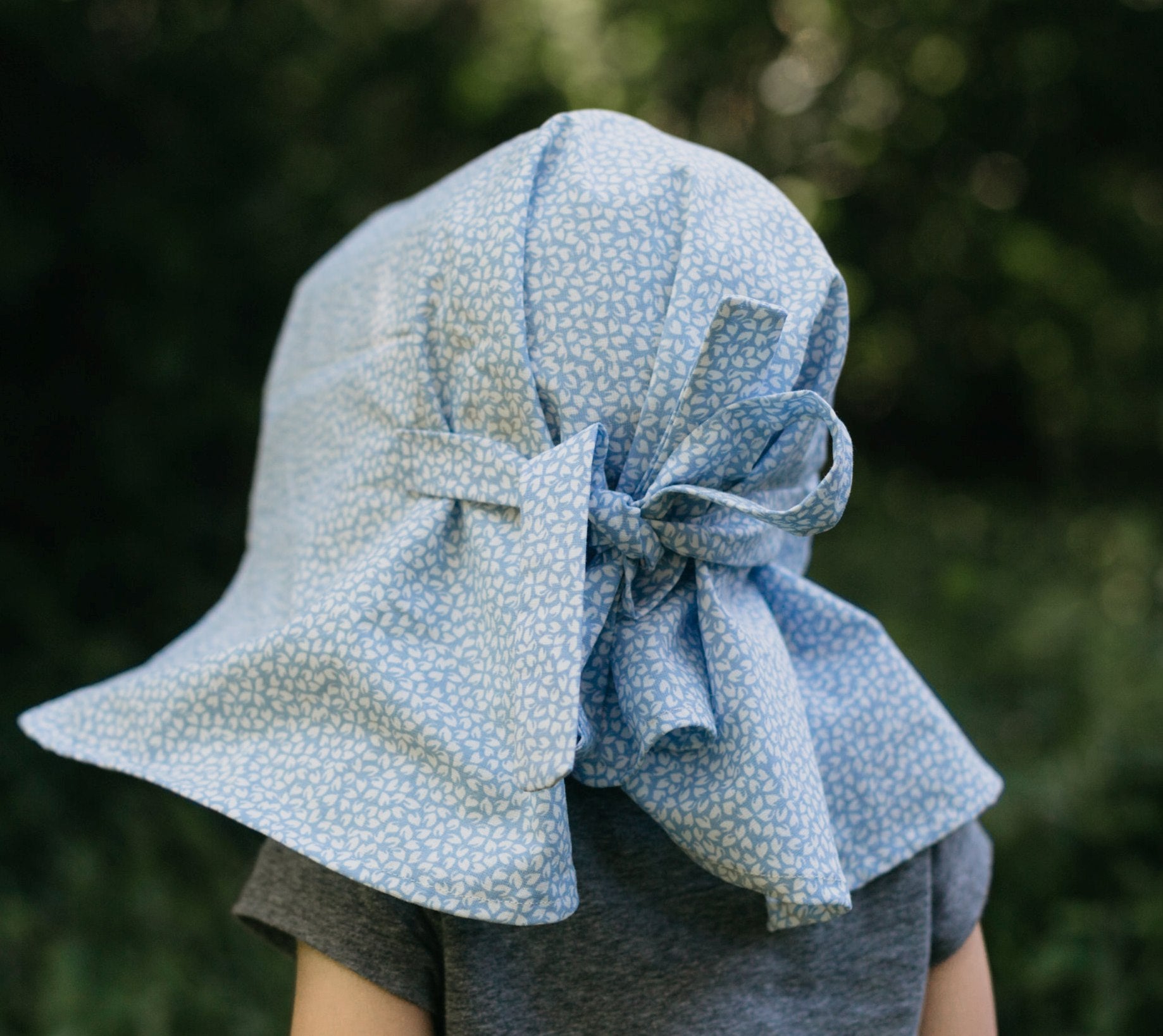 Back view of little girl surrounded by greenery wearing 213 sunbonnet.