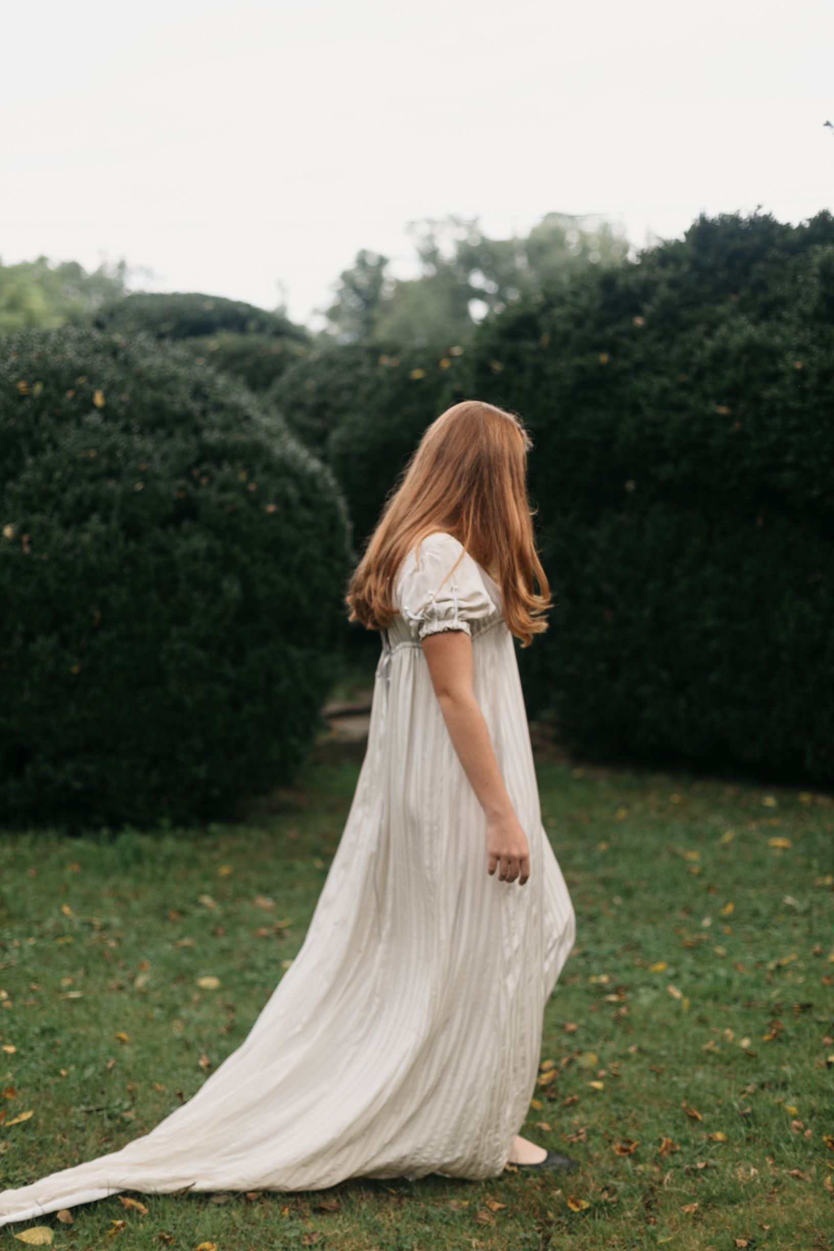 Side view of red head young woman standing surrounded greenery wearing 215 Empire Dress.