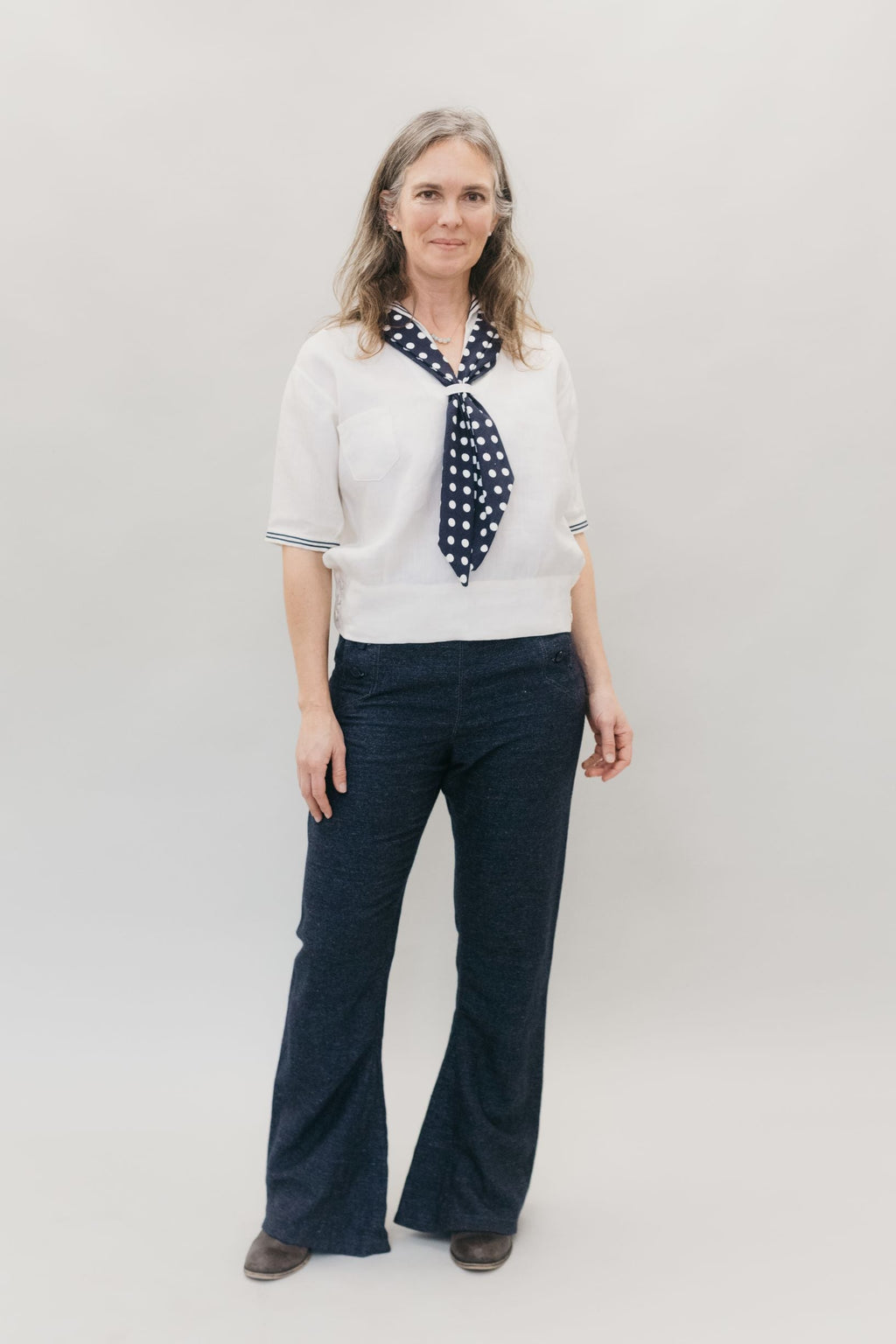 Woman smiling and standing in front of a white studio backdrop wearing 229 Sailor pants and 211 Two middies with scarf.