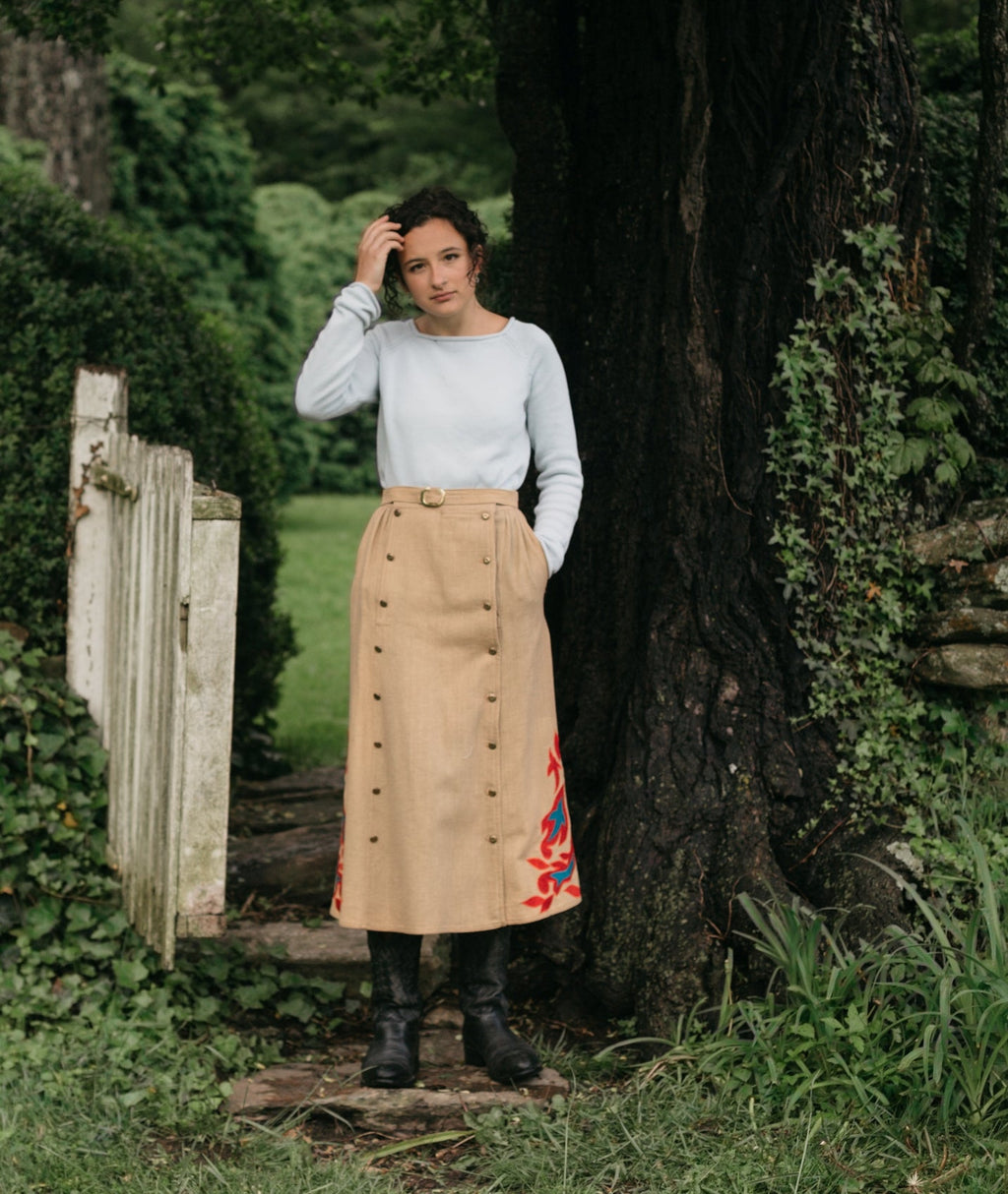 Woman standing in front of an open gate surrounded by greenery wearing the 231 Big Sky Riding Skirt buttoned to the left for a skirt effect.