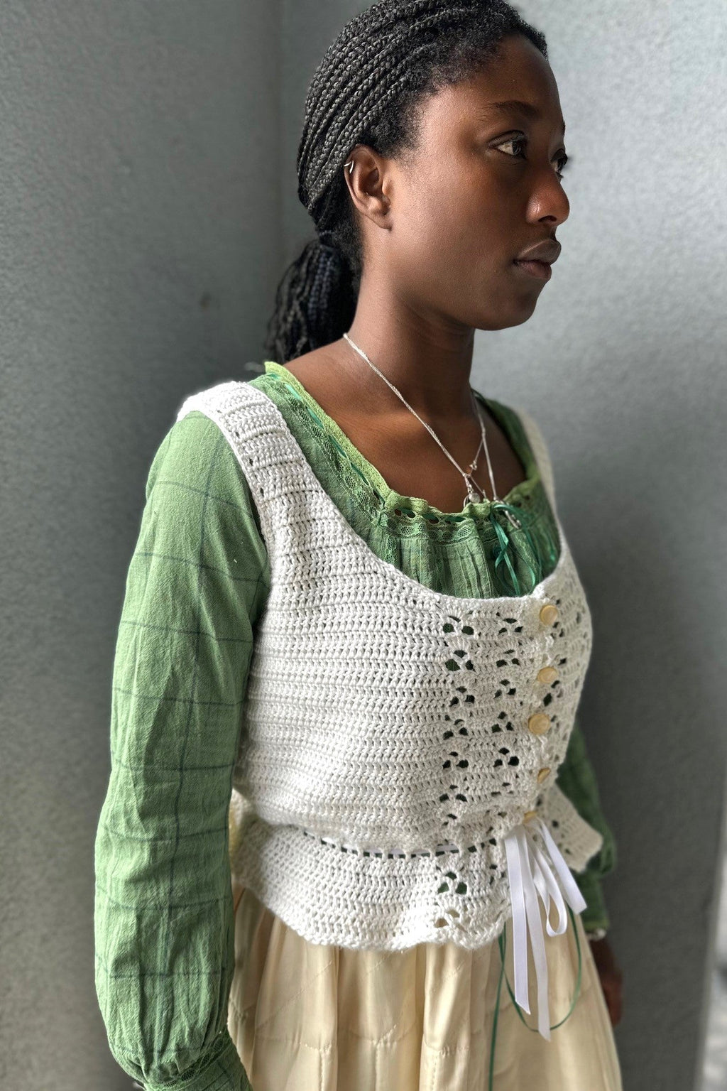 Black woman wearing a cream quilted skirt with a green blouse and white crocheted camisole on top - in front of a grey wall.