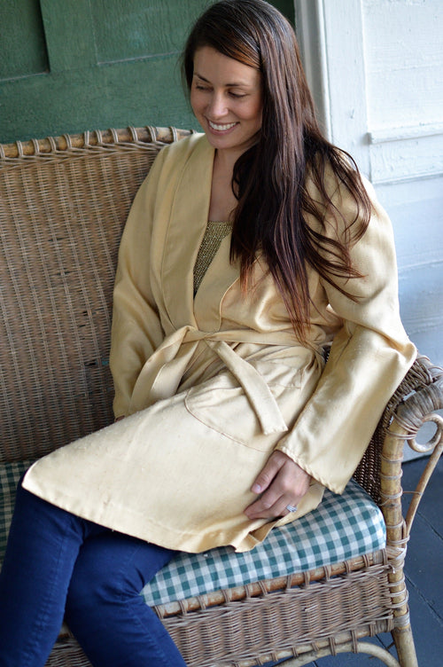 Brunette woman sitting on a woven chair legs crossed wearing 238 Le Smoking Jacket with sash wrapped around her waist.
