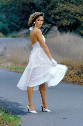 Woman wearing a white blonde bombshell dress on a road while twirling. She is wearing white kid gloves.