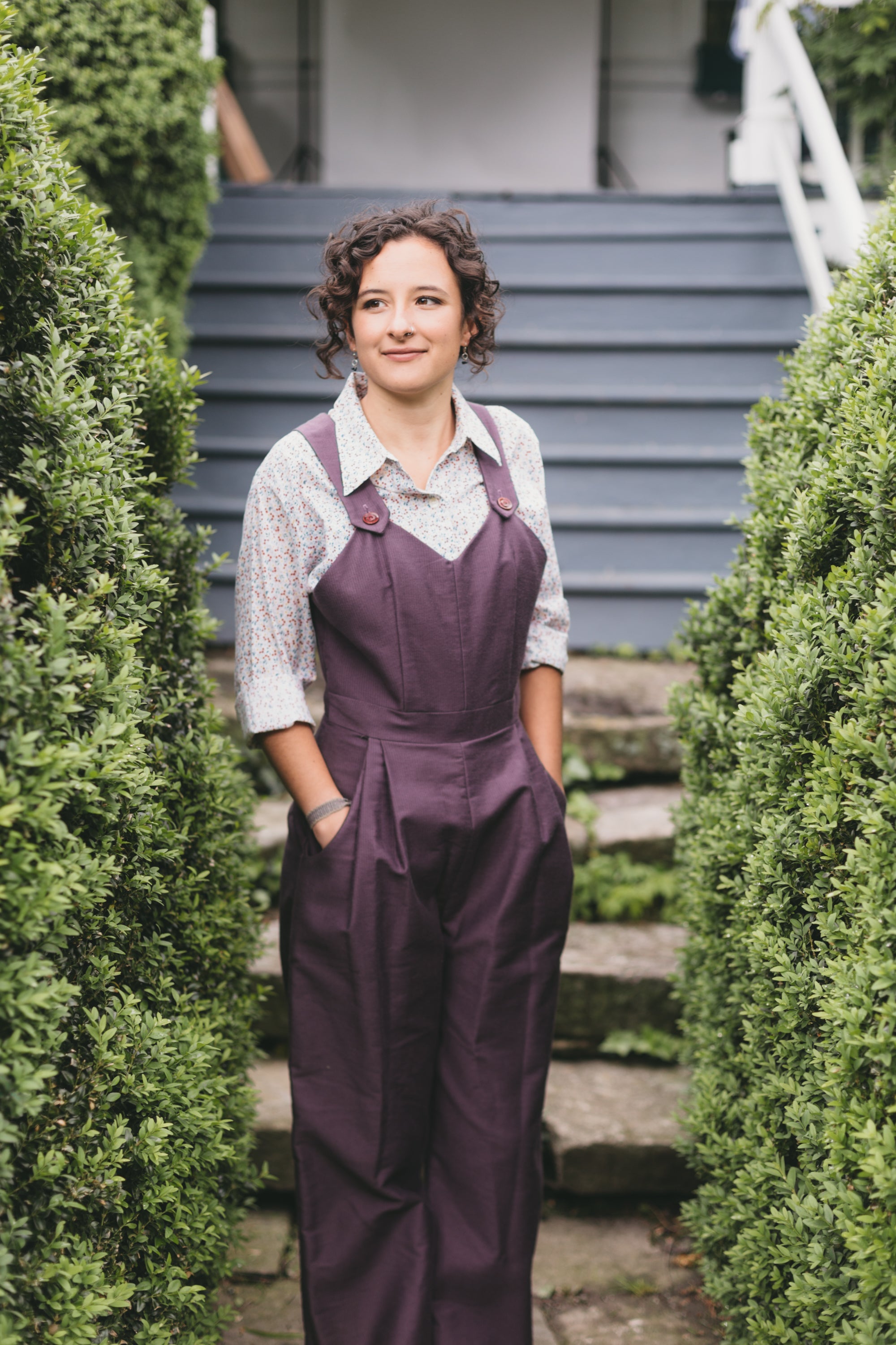 Young brunette white woman standing with hands in pockets of the 240 Rosie the Riveter overalls surrounded by greenery.