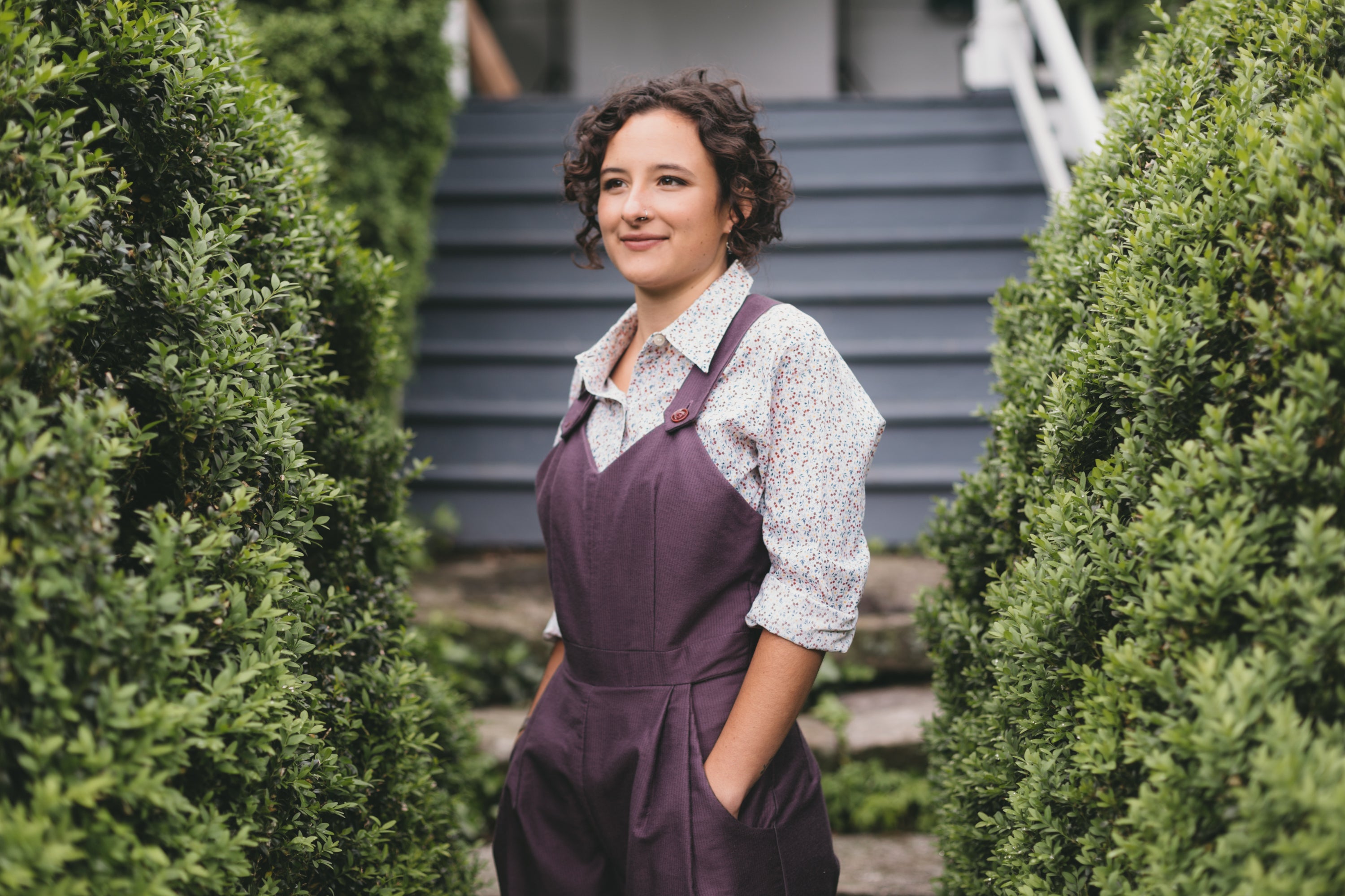 Young brunette woman turned slightly to the left wearing the Rosie the Riveter overalls with hands in the pockets surrounded by greenery.