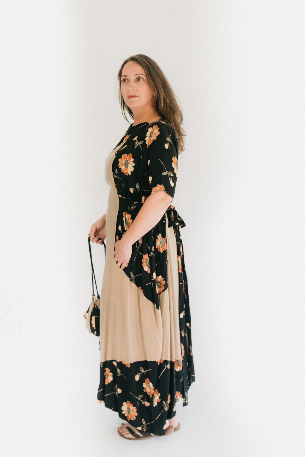 A brunette woman standing in front of a white studio backdrop, turned to the side wearing #261 Paris Promenade Dress, holding a handbag.