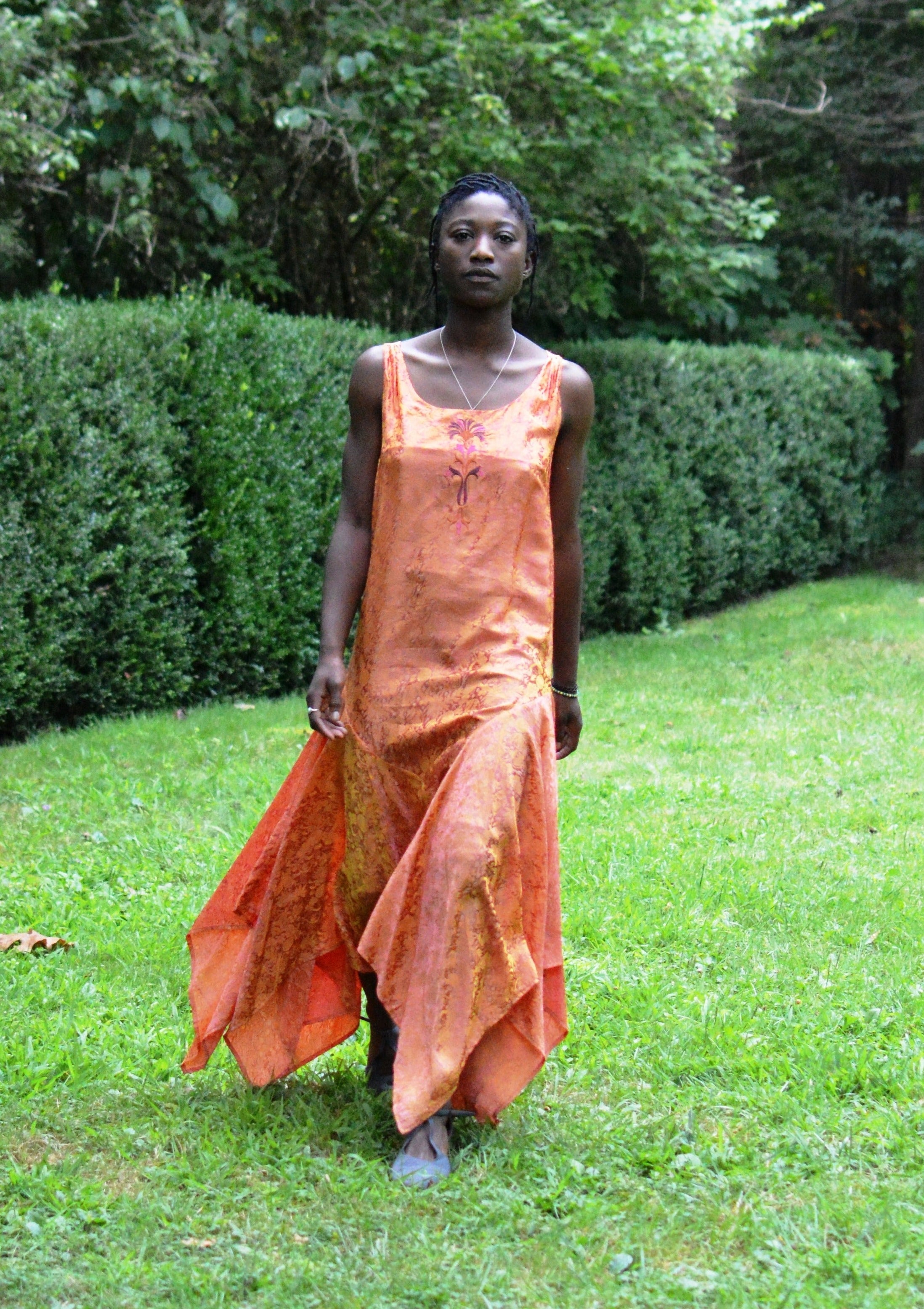 Young African woman standing surrounded by greenery wearing #264 Monte Carlo Dress in orange.