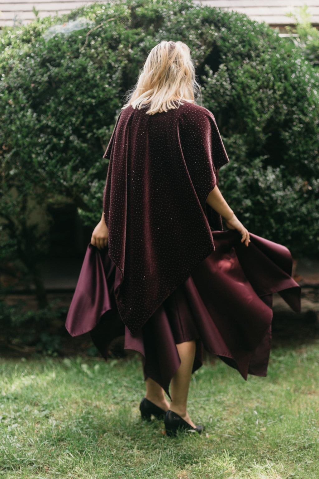 Back view of young blonde white woman standing surrounded by greenery wearing the Monte Carlo dress and V-Neck Tunic in maroon.