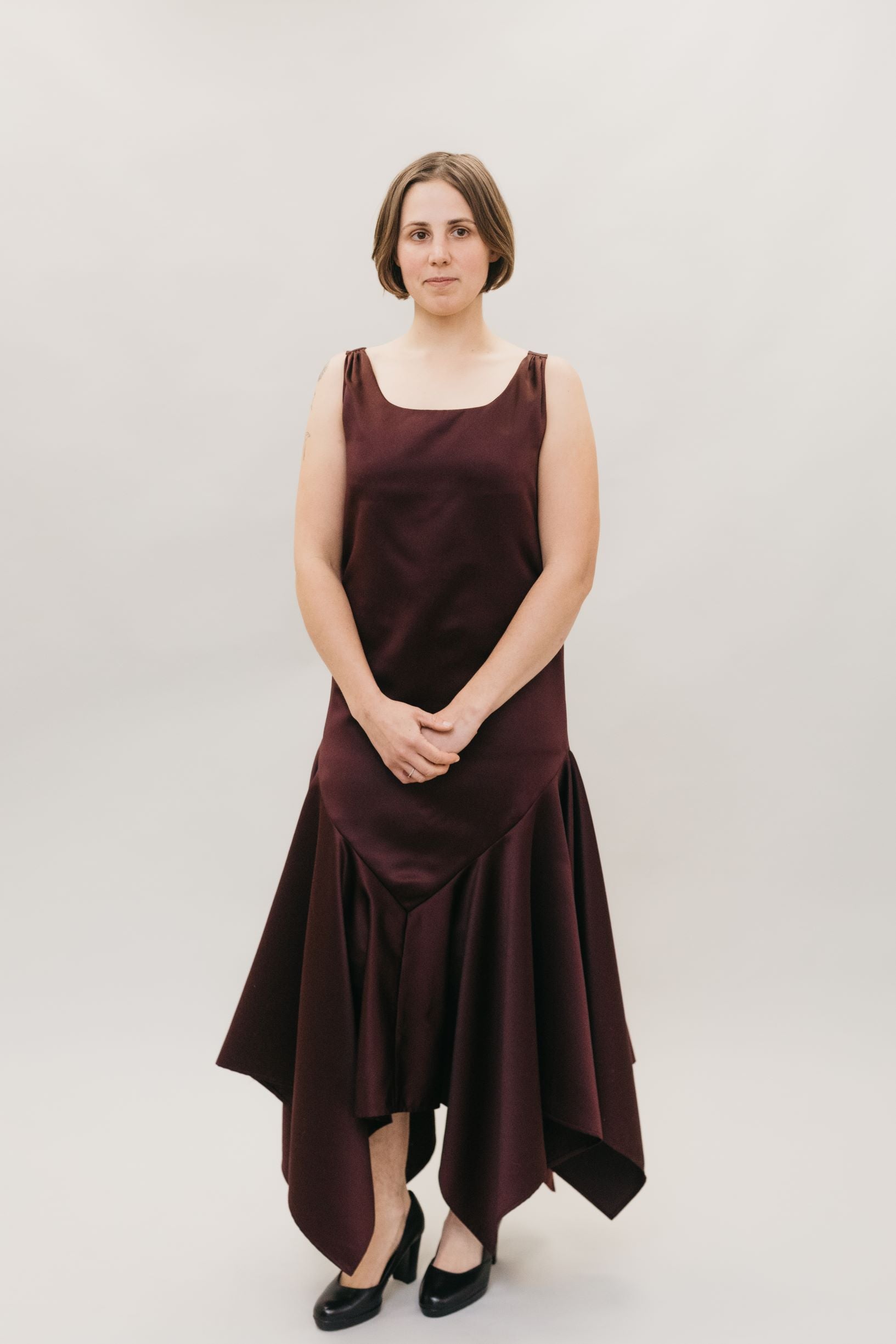 Young Burnett White woman with neck length hair, standing in front of a studio white backdrop wearing the #264 Monte Carlo Dress in maroon., with black pumps.