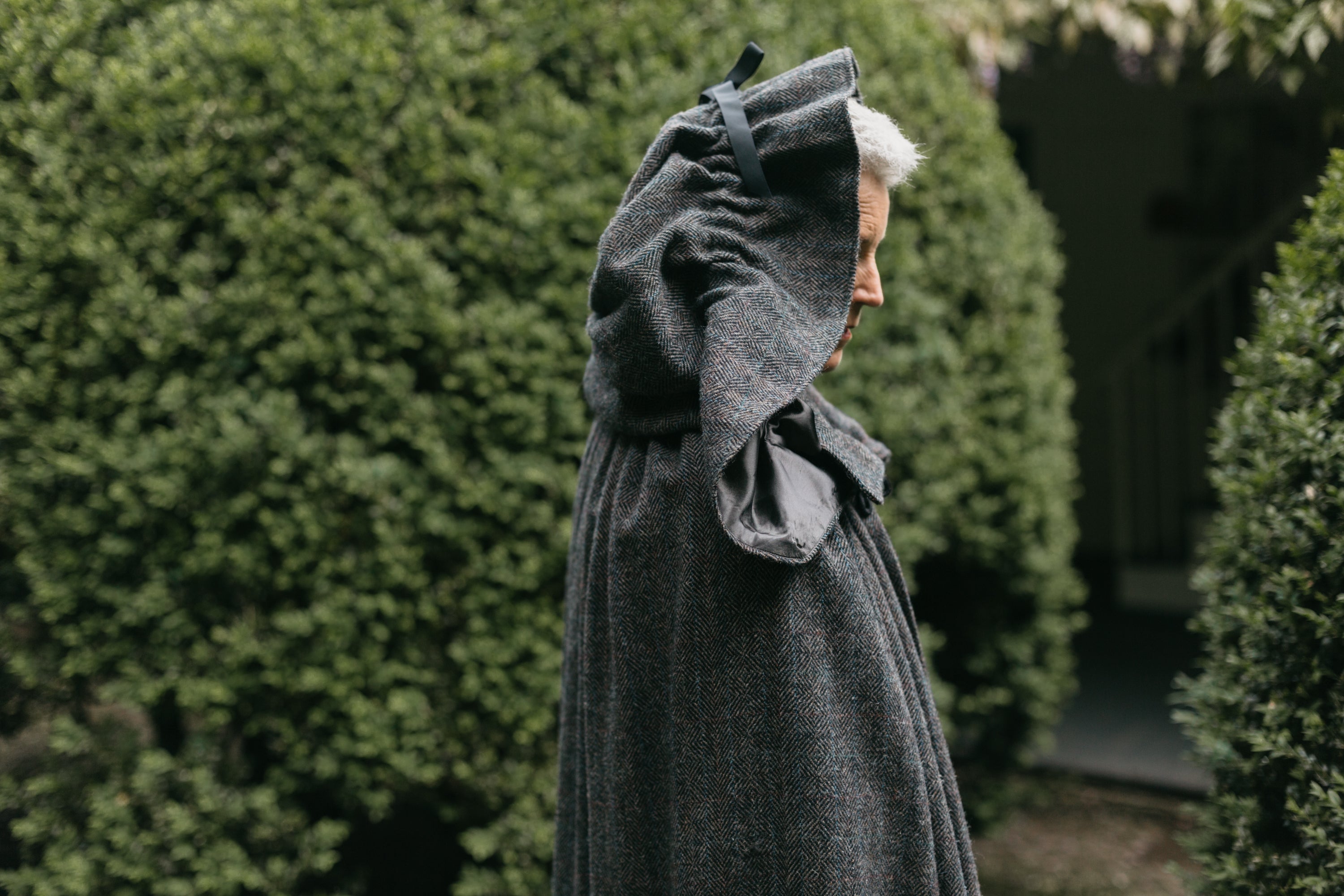 Close up of older white woman standing surrounded by greenery wearing 207 Kinsale Cloak with hood up.