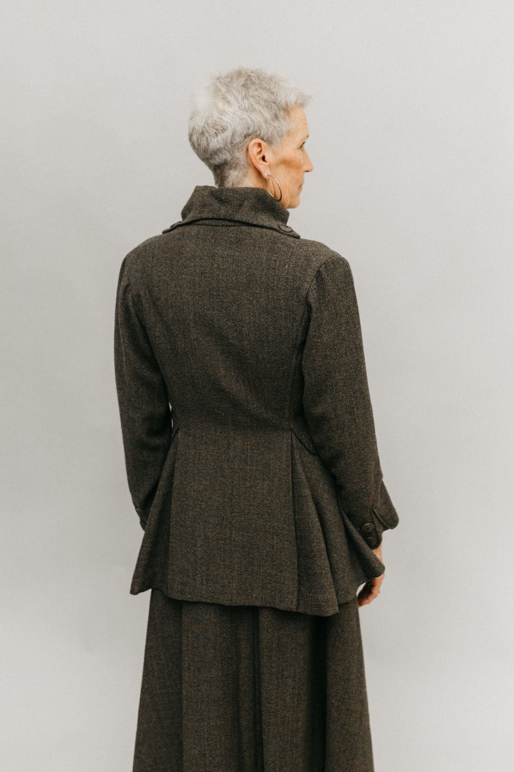Older white woman standing in font of a white studio backdrop, wearing the traveling suit back facing the audience showing the flared below the hip peplum and the high stand collar