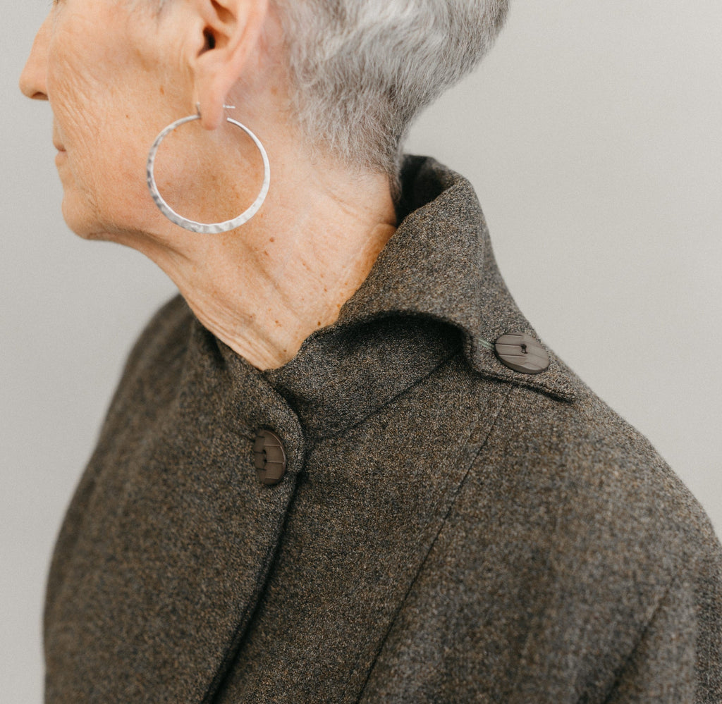 Older white woman wearing sliver hooped earnings ,close up of her left shoulder and neck wearing the Traveling Jacket buttoned on the left side on the neck, with a contrast over collar