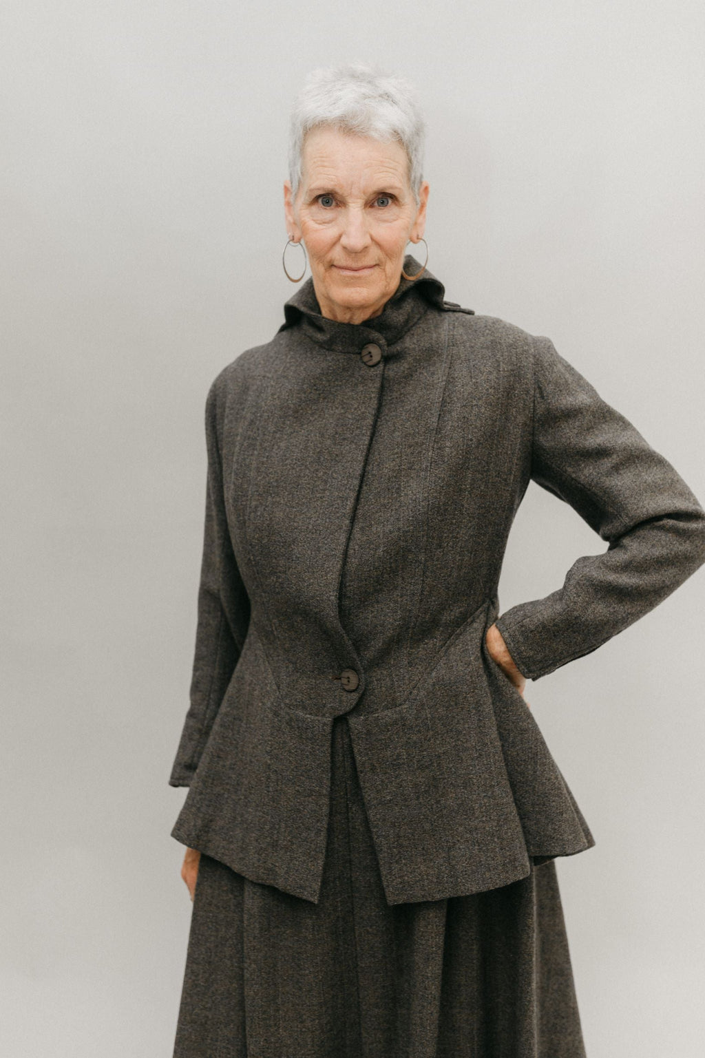 Older white woman with gray hair with her left hand on her hip, standing in front of a white studio backdrop wearing the traveling suit buttoned