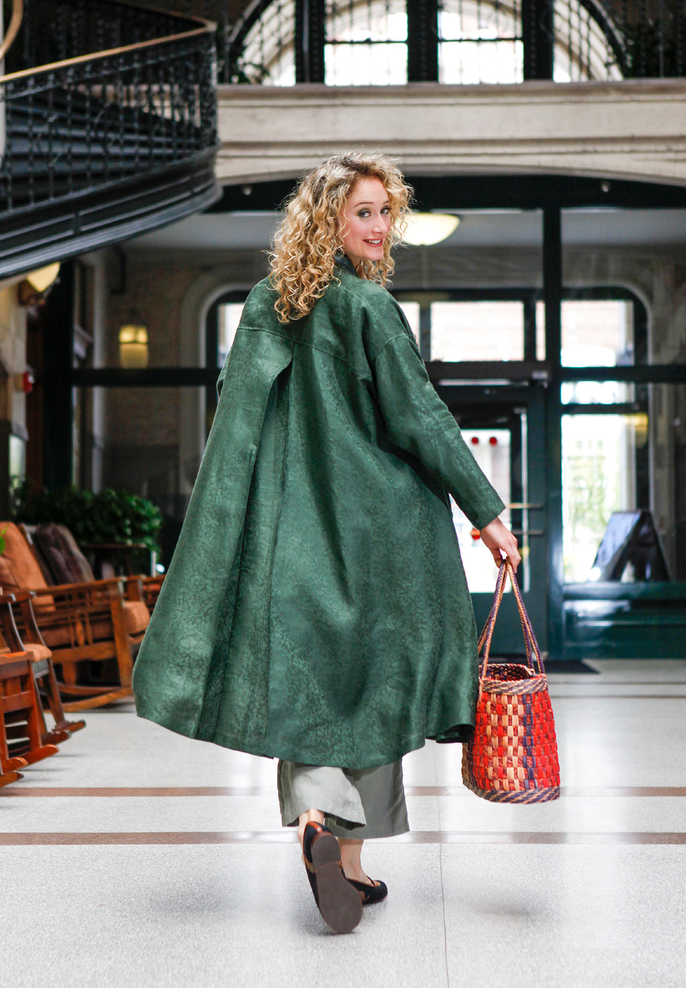 Back view of a blonde white woman looking over her shoulder holding a woven purse wearing 254 Swing Coat open.